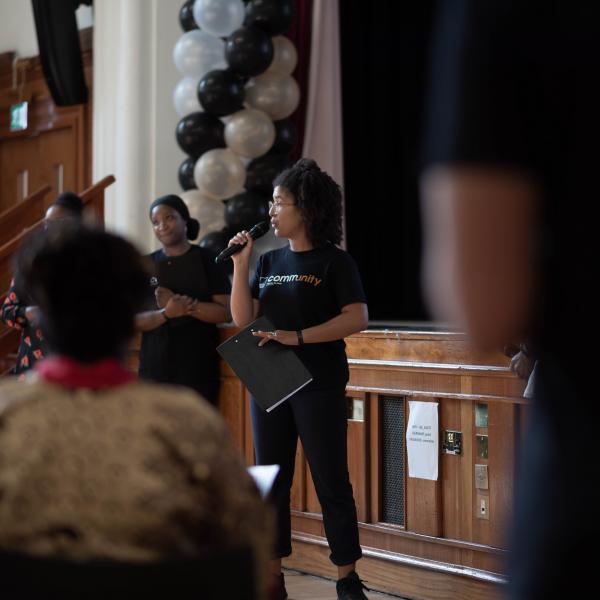 Woman leading community event with microphone against backdrop of Black Thrive branding and black and white balloons