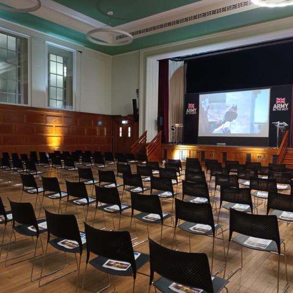 Assembly Hall with rows of chairs facing stage and screen, with red velvet curtains and uplighters around the room